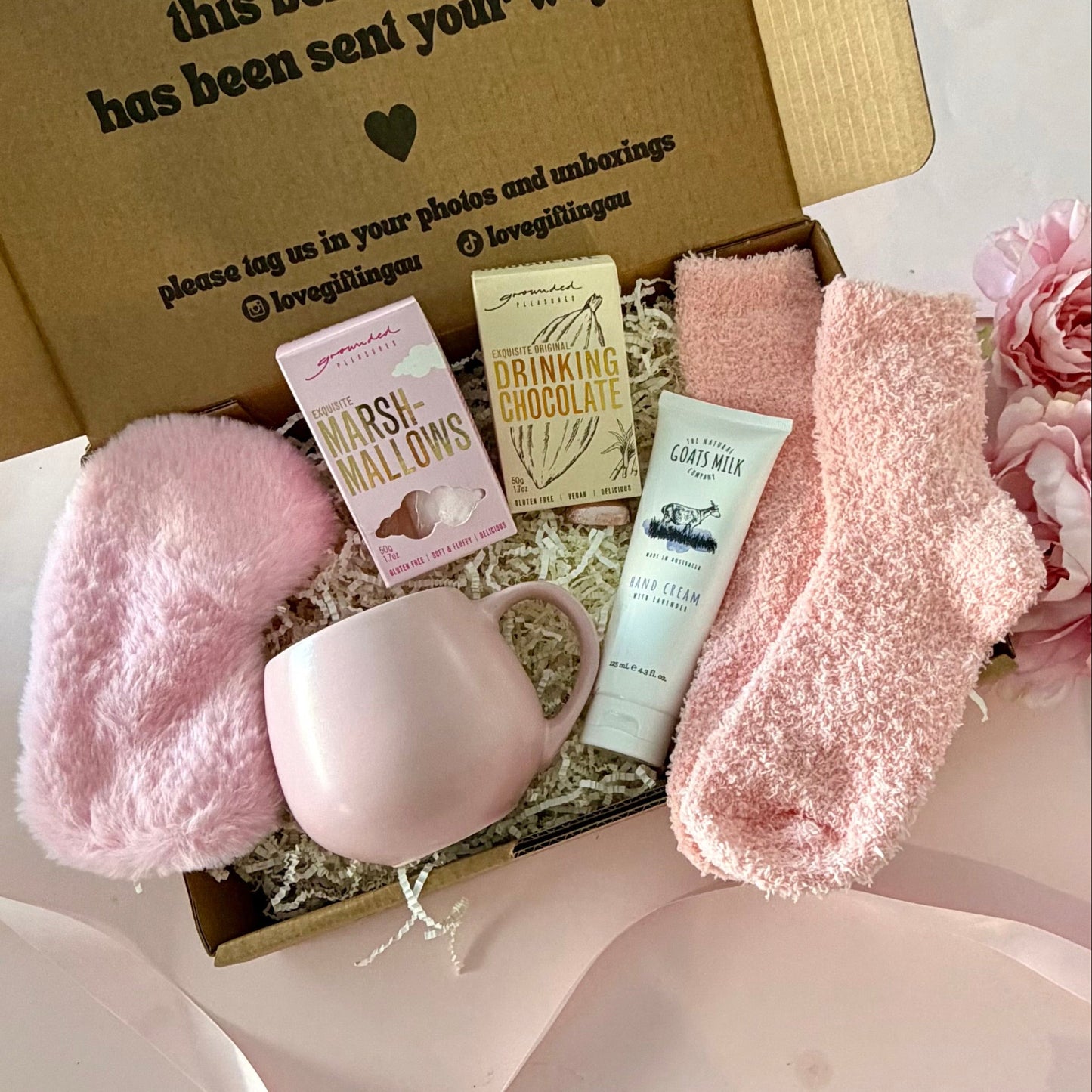 A curated comfort winter gift box featuring a pink mug, fluffy pink socks, gourmet marshmallow box, Grounded Pleasures hot chocolate box, mini chocolate heart, pink fluffy sleep mask and a lavender tube of hand cream 