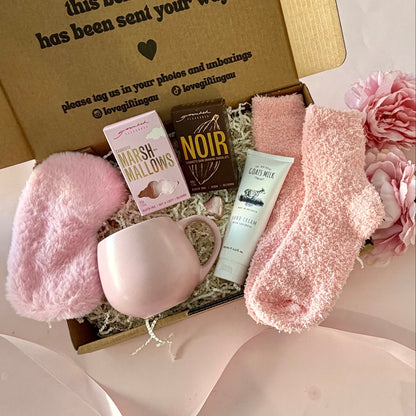 A curated comfort winter gift box featuring a pink mug, fluffy pink socks, gourmet marshmallow box, Grounded Pleasures hot chocolate box, mini chocolate heart, pink fluffy sleep mask and a lavender tube of hand cream  