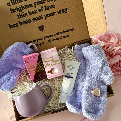 A curated comfort winter gift box featuring a purple mug, fluffy purple socks, gourmet marshmallow box, Grounded Pleasures hot chocolate box, mini chocolate heart, purple fluffy sleep mask and a lavender tube of hand cream  