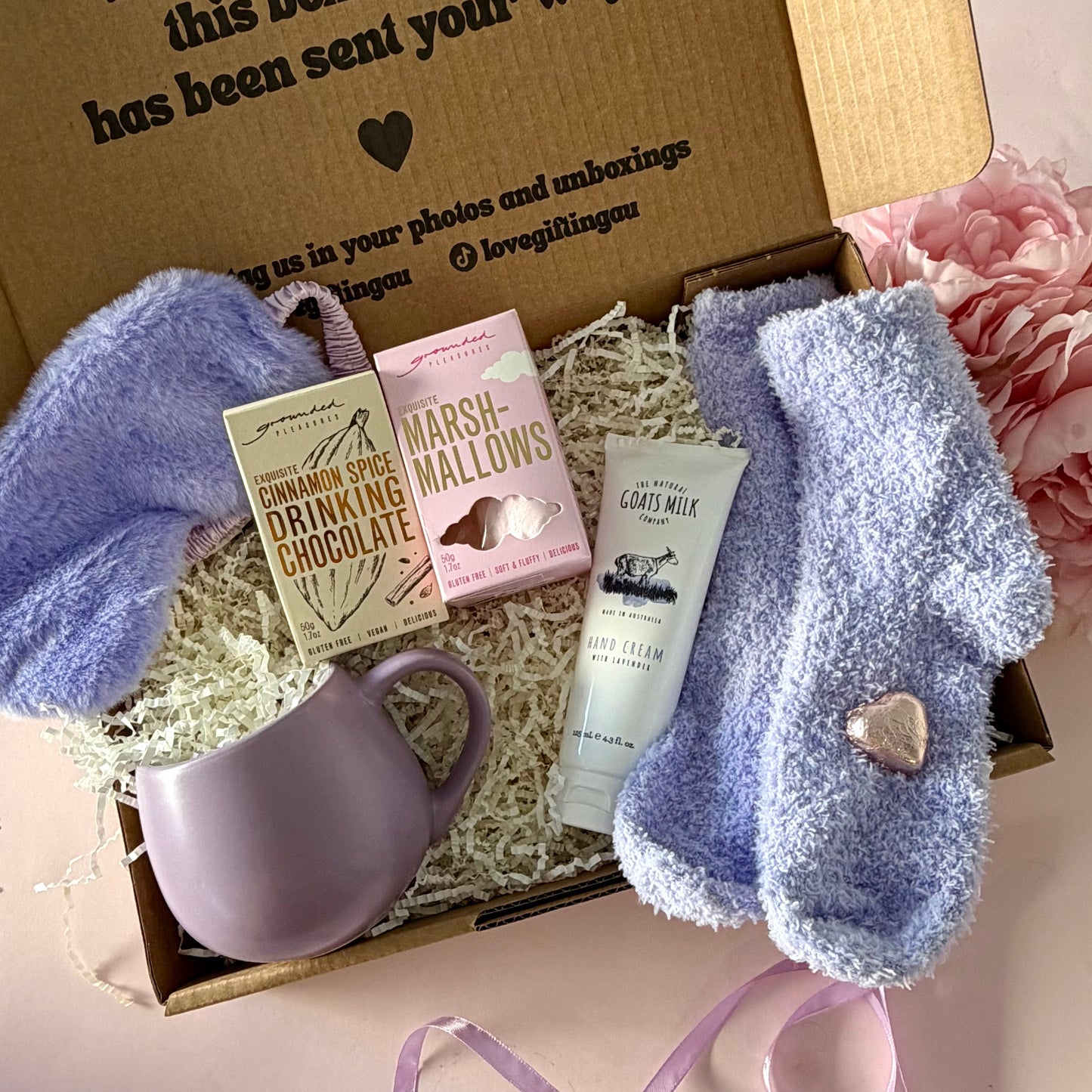 A curated comfort winter gift box featuring a purple mug, fluffy purple socks, gourmet marshmallow box, Grounded Pleasures hot chocolate box, mini chocolate heart, purple fluffy sleep mask and a lavender tube of hand cream  