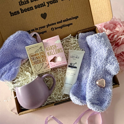 A curated comfort winter gift box featuring a purple mug, fluffy purple socks, gourmet marshmallow box, Grounded Pleasures hot chocolate box, mini chocolate heart, purple fluffy sleep mask and a lavender tube of hand cream  