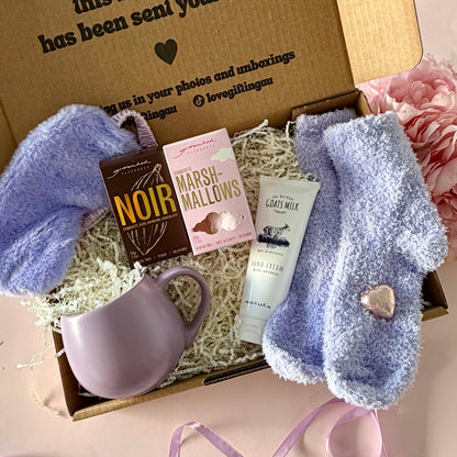 A curated comfort winter gift box featuring a purple mug, fluffy purple socks, gourmet marshmallow box, Grounded Pleasures hot chocolate box, mini chocolate heart, purple fluffy sleep mask and a lavender tube of hand cream  