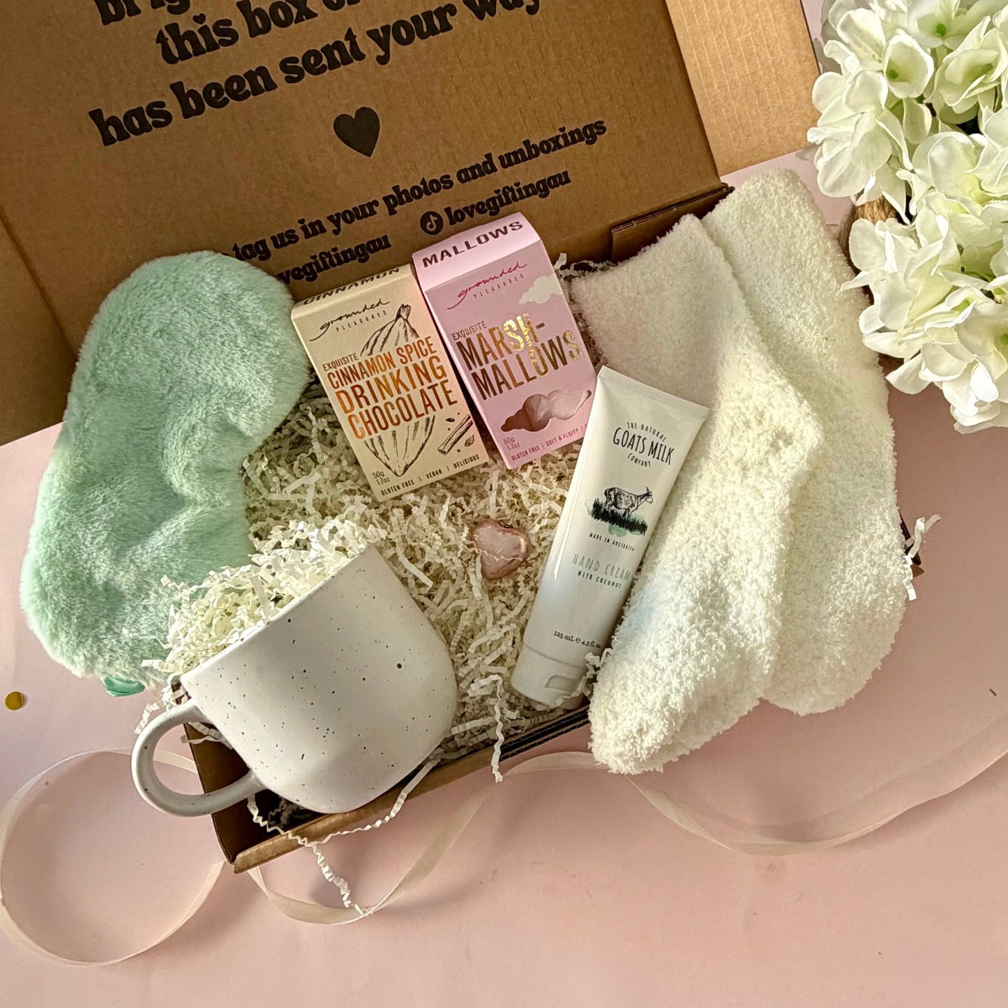 A curated comfort winter gift box featuring a cream speckled mug, warm fluffy cream socks, gourmet marshmallow box, Grounded Pleasures hot chocolate box, mini chocolate heart, pastel green fluffy sleep mask and a tube of hand cream with coconut.  