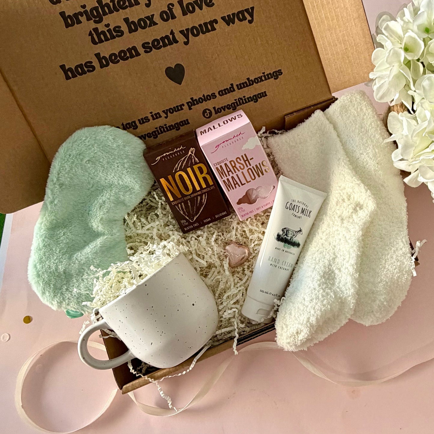 A curated comfort winter gift box featuring a cream speckled mug, warm fluffy cream socks, gourmet marshmallow box, Grounded Pleasures hot chocolate box, mini chocolate heart, pastel green fluffy sleep mask and a tube of hand cream with coconut.  