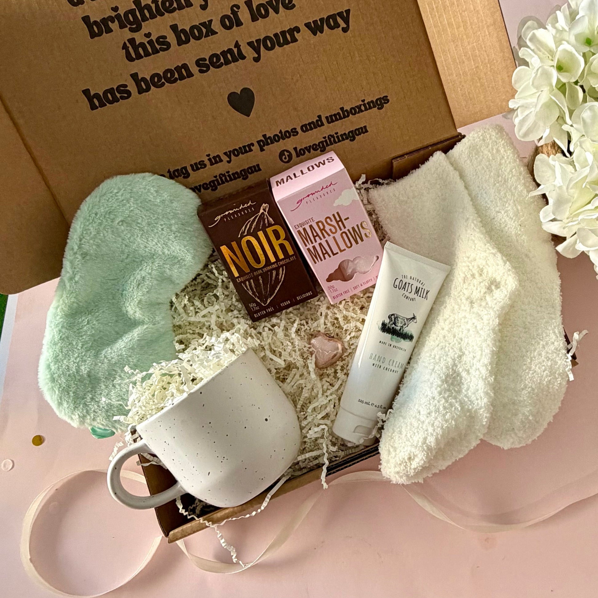 A curated comfort winter gift box featuring a cream speckled mug, warm fluffy cream socks, gourmet marshmallow box, Grounded Pleasures hot chocolate box, mini chocolate heart, pastel green fluffy sleep mask and a tube of hand cream with coconut.  