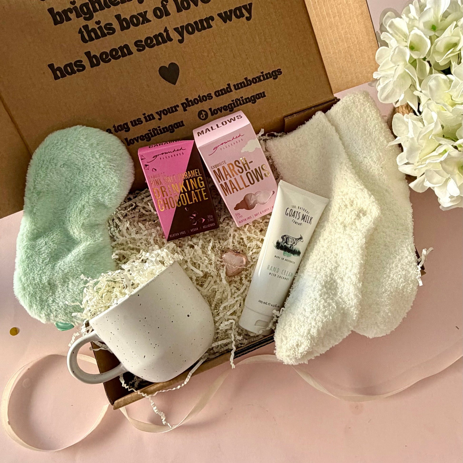 A curated comfort winter gift box featuring a cream speckled mug, warm fluffy cream socks, gourmet marshmallow box, Grounded Pleasures hot chocolate box, mini chocolate heart, pastel green fluffy sleep mask and a tube of hand cream with coconut.  