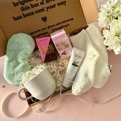 A curated comfort winter gift box featuring a cream speckled mug, warm fluffy cream socks, gourmet marshmallow box, Grounded Pleasures hot chocolate box, mini chocolate heart, pastel green fluffy sleep mask and a tube of hand cream with coconut.  