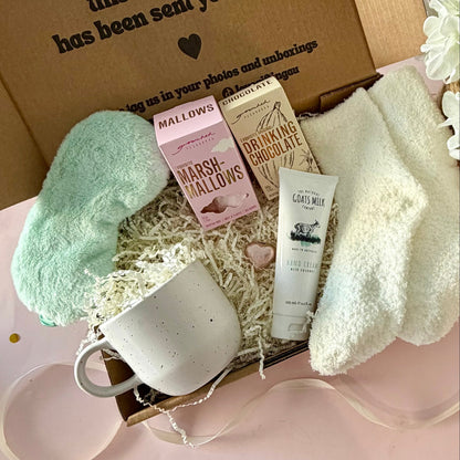 A curated comfort winter gift box featuring a cream speckled mug, warm fluffy cream socks, gourmet marshmallow box, Grounded Pleasures hot chocolate box, mini chocolate heart, pastel green fluffy sleep mask and a tube of hand cream with coconut.  