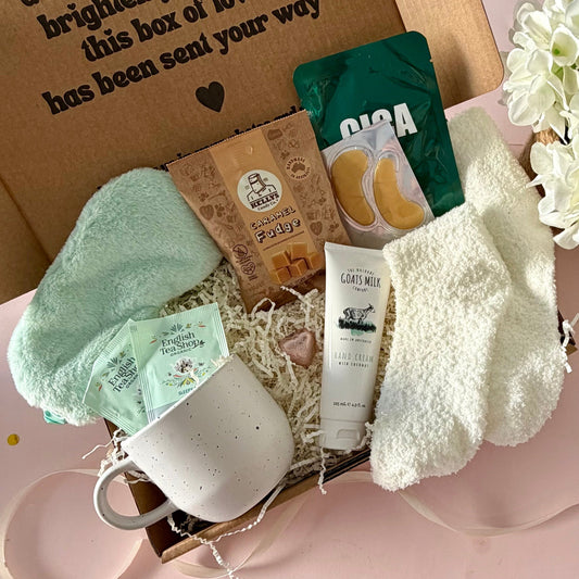 A curated comfort winter gift box featuring a cream speckled mug, Sleepy me teabags, Kellys Candy Co Fudge, warm fluffy cream socks, mini chocolate heart, pastel green fluffy sleep mask and a tube of hand cream with coconut.  