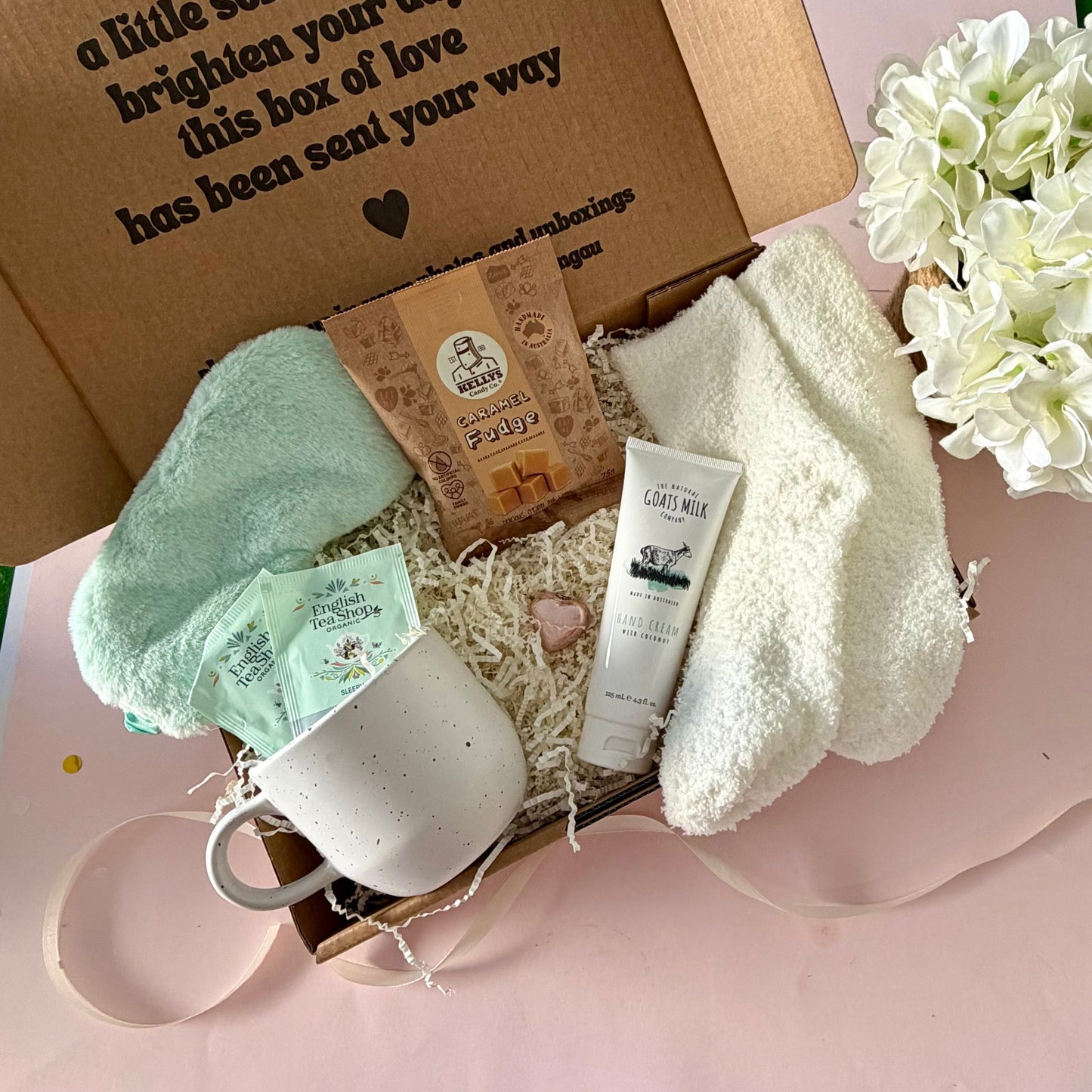 A curated comfort winter gift box featuring a cream speckled mug, Sleepy me teabags, Kellys Candy Co Fudge, warm fluffy cream socks, mini chocolate heart, pastel green fluffy sleep mask , lapcos facemask sheet and a tube of hand cream with coconut. 