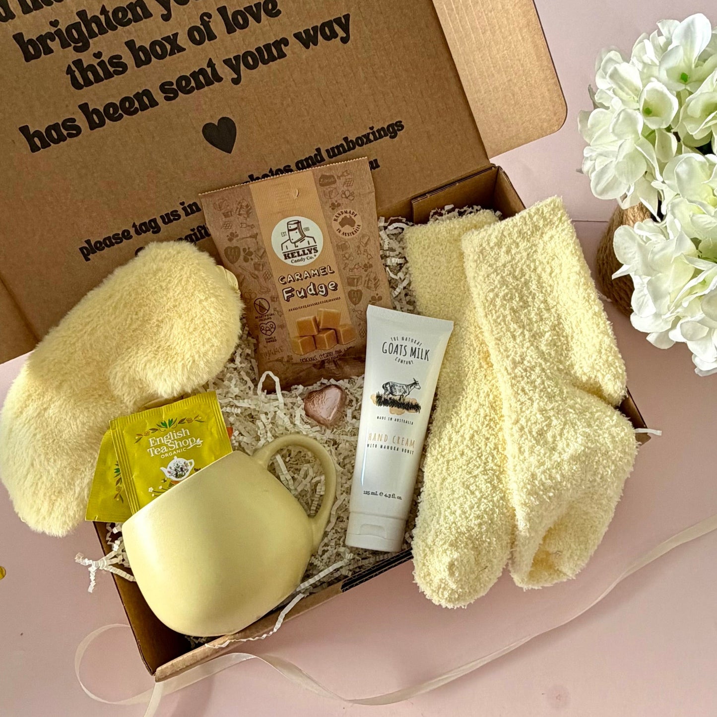 A curated comfort winter gift box featuring a yellow mug, lemongrass teabags, Kellys Candy Co Fudge, warm fluffy yellow socks, mini chocolate heart, yellow fluffy sleep mask and a tube of hand cream.  