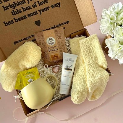 A curated comfort winter gift box featuring a yellow mug, lemongrass teabags, Kellys Candy Co Fudge, warm fluffy yellow socks, mini chocolate heart, yellow fluffy sleep mask and a tube of hand cream.  
