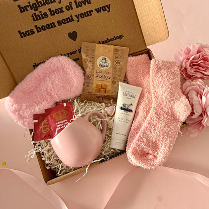 A curated comfort winter gift box featuring a pink cuddle mug, teabags, Kellys Candy Co Fudge, warm fluffy pink socks, mini chocolate heart, pink fluffy sleep mask and a tube of hand cream.  