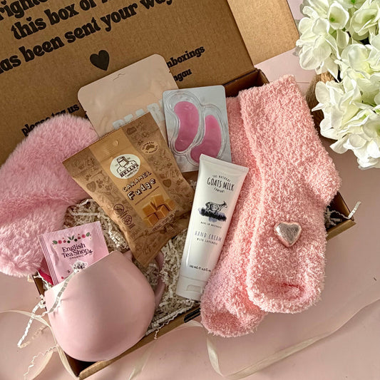 A curated comfort winter gift box featuring a pink cuddle mug, teabags, Kellys Candy Co Fudge, warm fluffy pink socks, mini chocolate heart, pink fluffy sleep mask and a tube of hand cream.  Lapcos Pearl Facemask sheet and pink under eye masks.