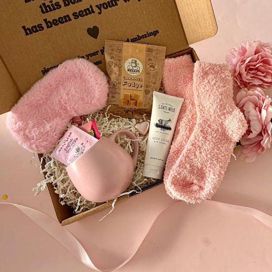 A curated comfort winter gift box featuring a pink cuddle mug, teabags, Kellys Candy Co Fudge, warm fluffy pink socks, mini chocolate heart, pink fluffy sleep mask and a tube of hand cream.  