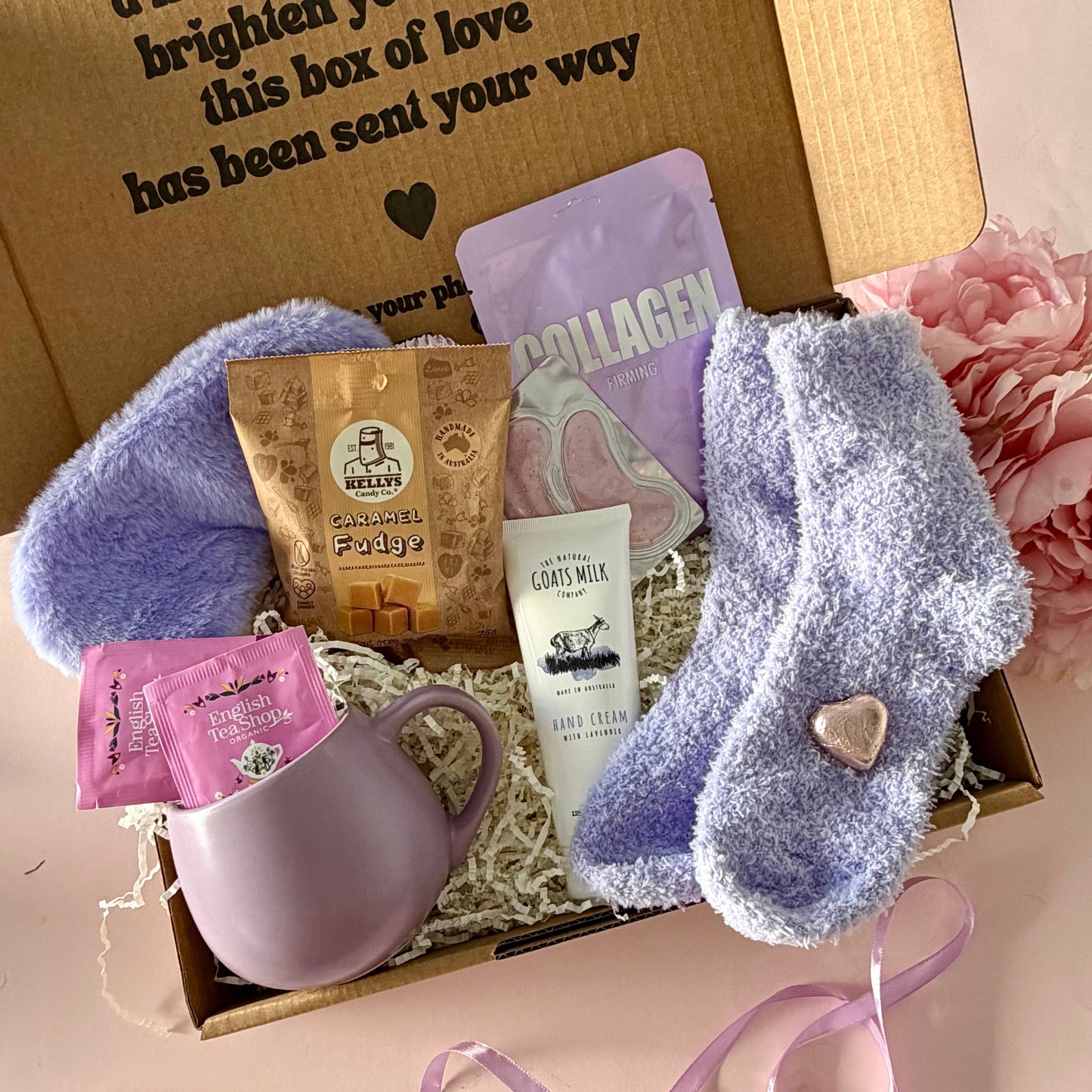 A curated comfort winter gift box featuring a purple cuddle mug, teabags, Kellys Candy Co Fudge, warm fluffy purple socks, mini chocolate heart, purple fluffy sleep mask and a tube of hand cream.  Lapcos collagen Facemask sheet and purple under eye masks.