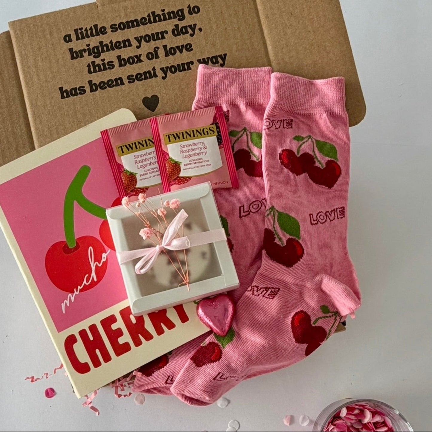 A cherry-themed gift box set on a white surface, featuring pink cherry-print socks, a pink 'muchacho CHERRY' notebook, Twinings tea packets, love shower steamer, and a chocolate heart