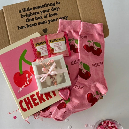 A cherry-themed gift box set on a white surface, featuring pink cherry-print socks, a pink 'muchacho CHERRY' notebook, Twinings tea packets, love shower steamer, and a chocolate heart