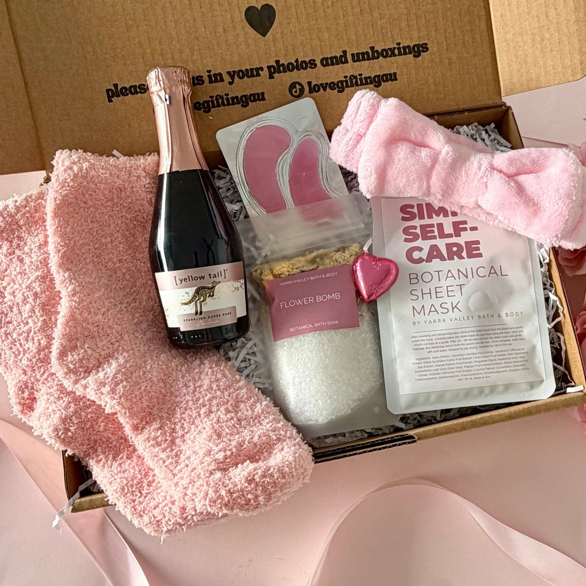 A curated self-care gift box shown from above, containing a pink towelling headband,  a small bottle of Rosé prosecco, bath salts,  Self-Care Botanical Sheet Mask. pink socks, under eye mask and a chocolate heart. The open cardboard box lid features printed text.