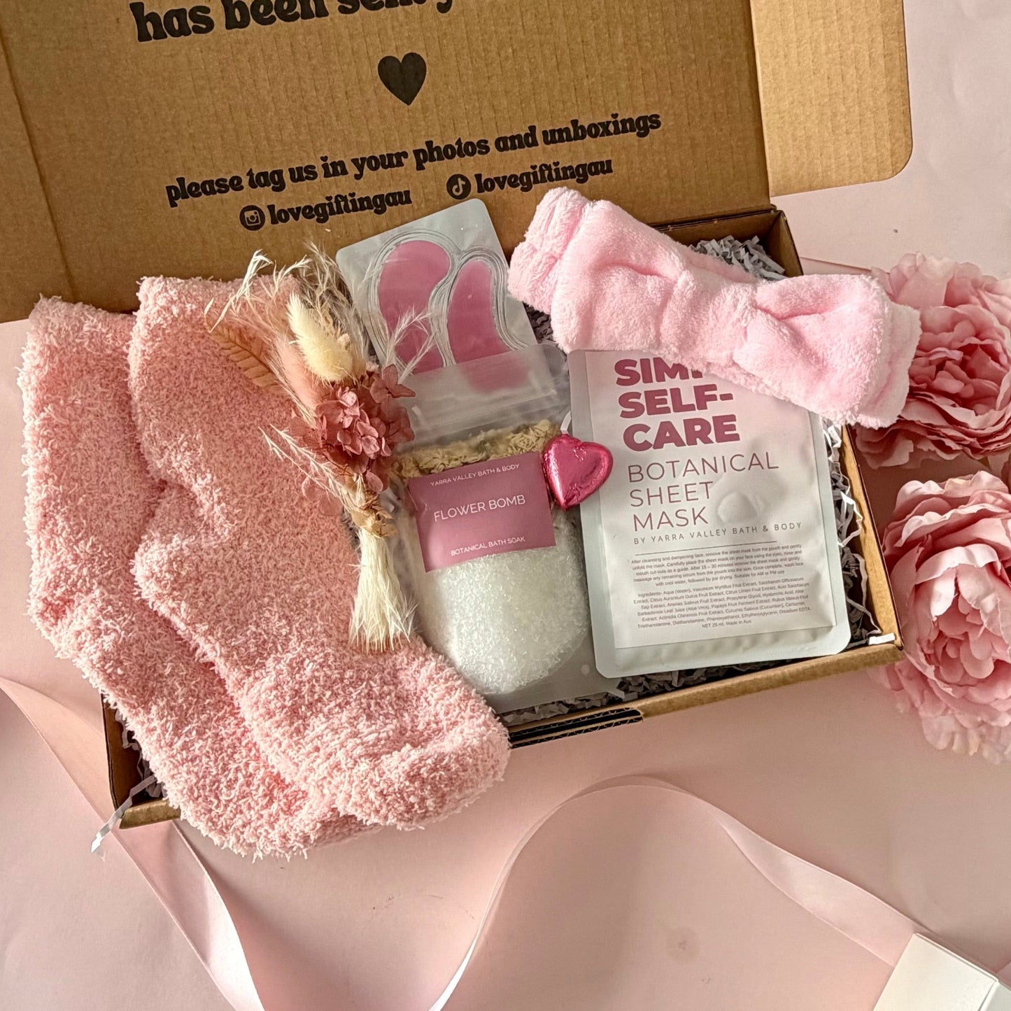 A pamper gift box filled with self-care items, including a pink towelling headband , a botanical sheet mask, bath salts, dried flower posy , under eye mask and a chocolate heart.