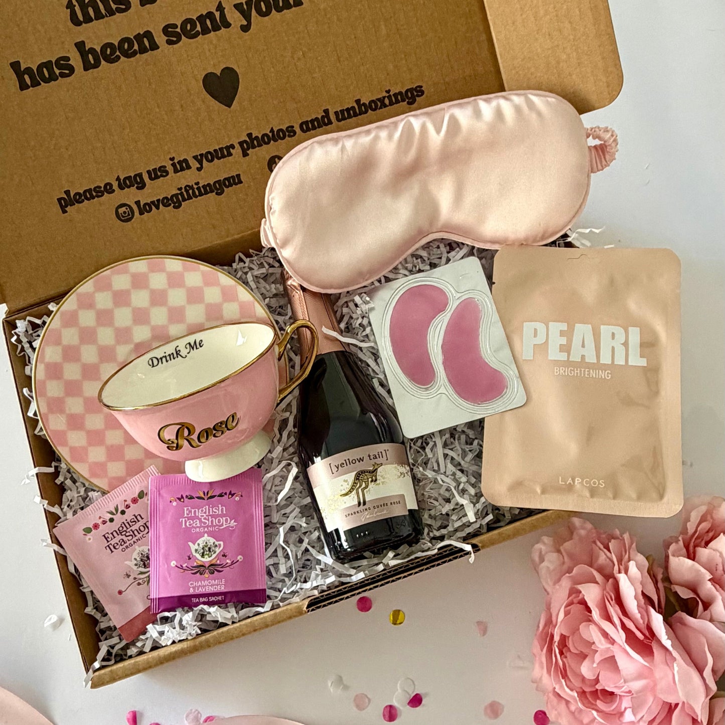 A curated gift box set displayed in an open kraft cardboard box on a white surface. The set includes a light pink satin sleep mask, a Rosé teacup,  a small Rosé prosecco bottle, Lapcos Facemask, under eye and teabags