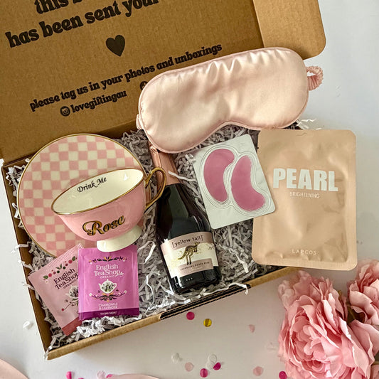 A curated gift box set displayed in an open kraft cardboard box on a white surface. The set includes a light pink satin sleep mask, a Rosé teacup,  a small Rosé prosecco bottle, Lapcos Facemask, under eye and teabags