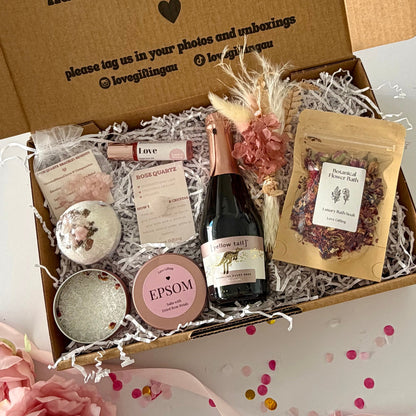 A curated  pamper self-care gift box set includes  a mini bottle of Rosé Prosecco , a rose quartz bath bomb, a tin of Epsom salts, a pouch of dried flowers, rose quartz hug, rose quartx bracelet and Love essential oil roller