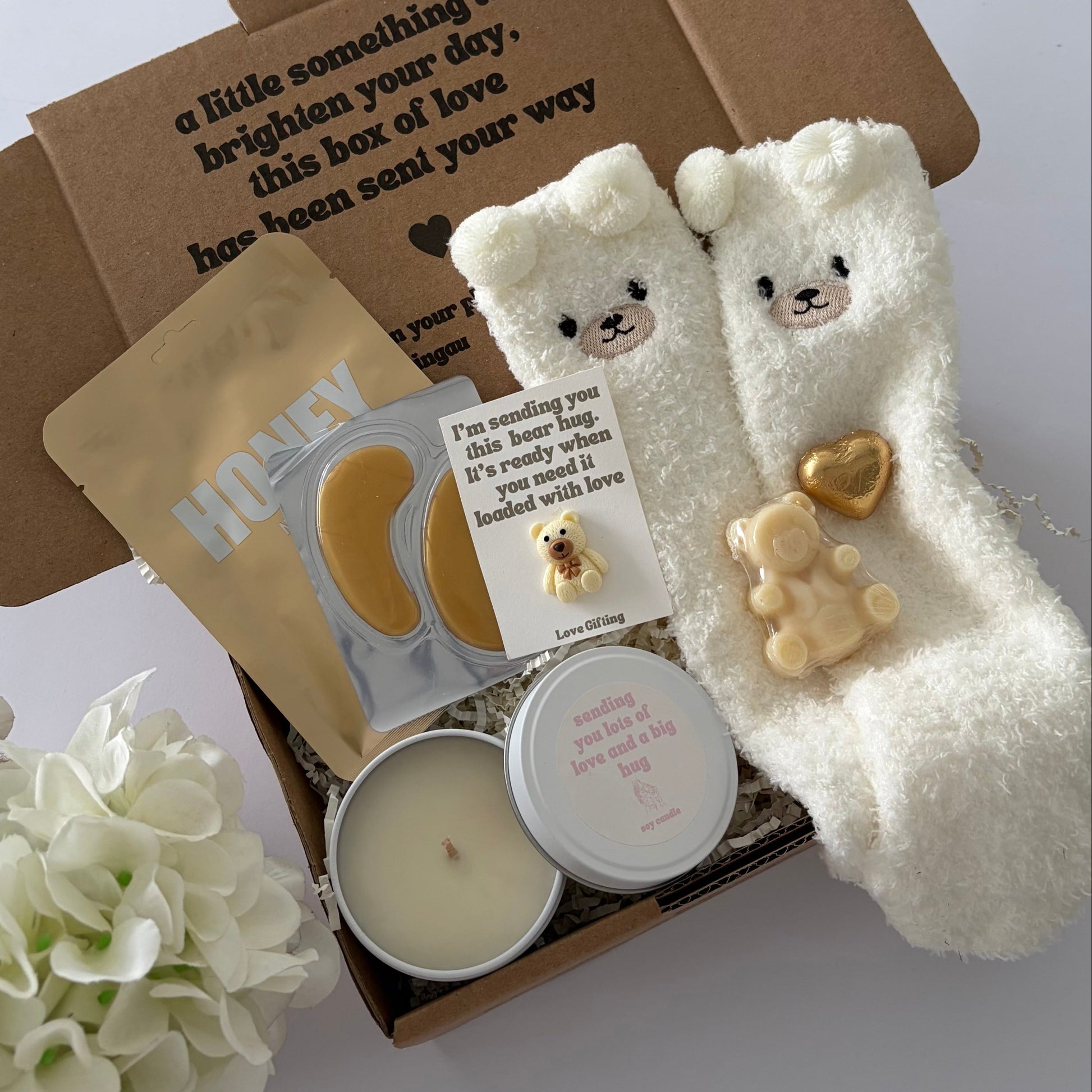 A curated gift box set containing a pair of fluffy white bear socks , Lapcos honey face mask sheet, gold, under eye,  a candle tin, bear soap, pocket hug and choc heart