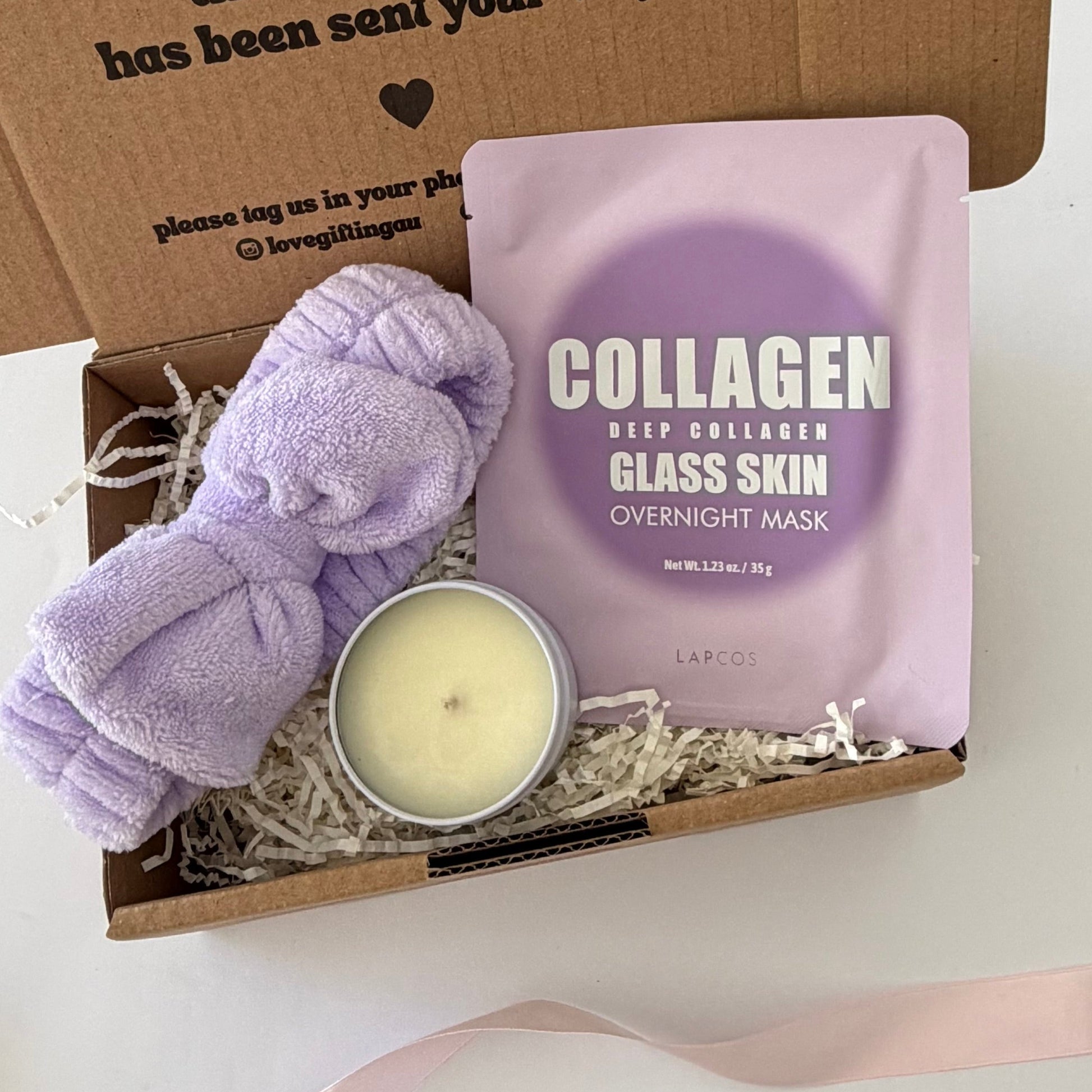 A gift box set containing a lavender plush towel,ling headband a small lavender glass jar candle, and a pink packet of 'COLLAGEN DEEP COLLAGEN GLASS SKIN OVERNIGHT MASK' by LAPCO, arranged in a brown cardboard box with shredded paper filler.