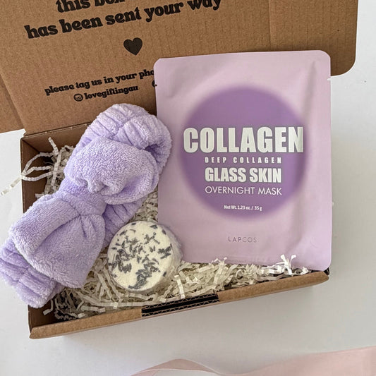 A gift box set featuring a lavender plush towel, a lavender collagen glass skin overnight mask pouch, and a lavender soap bar nestled in white paper shred inside a brown kraft box. The box lid has printed text, and a pink ribbon is draped in the foreground.