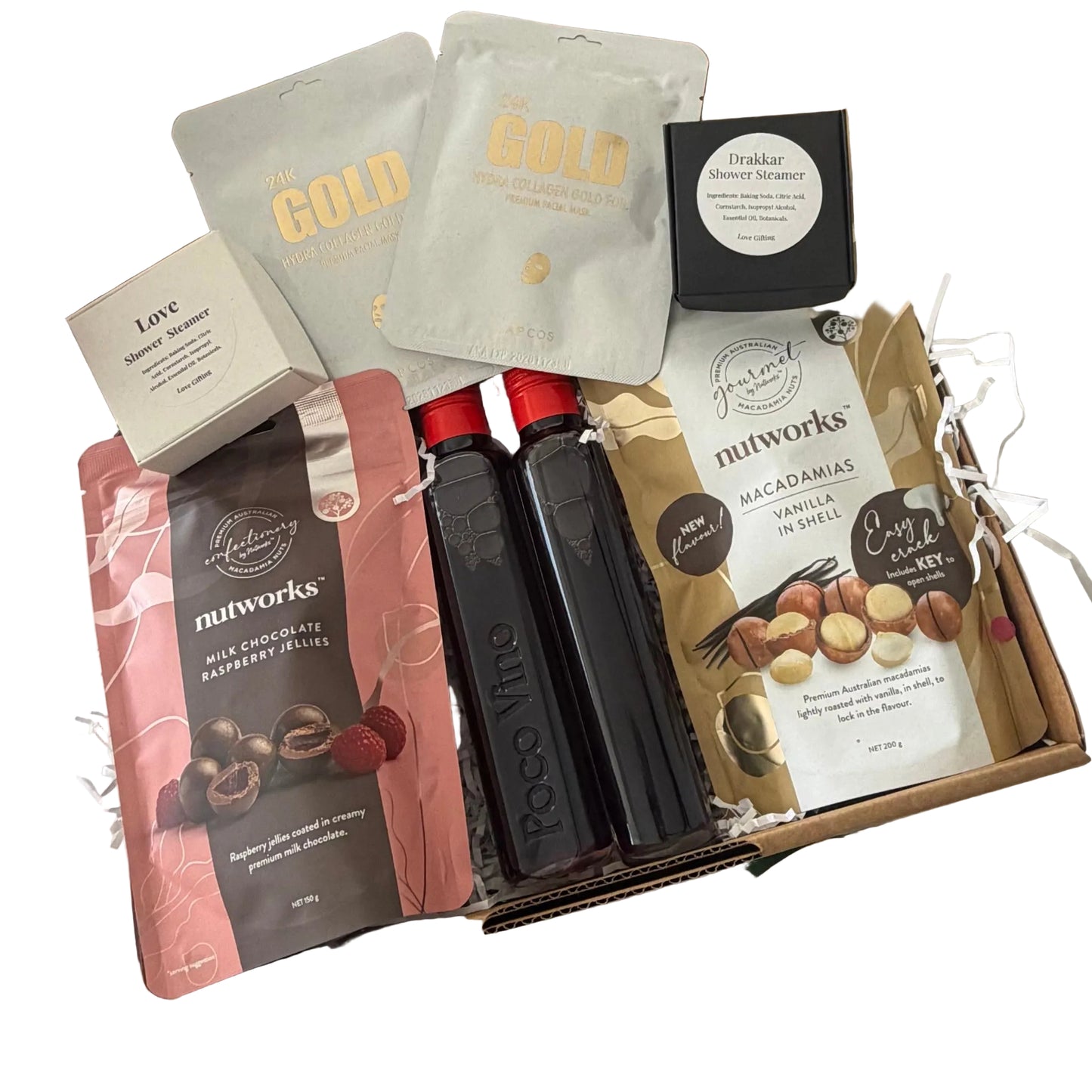 A curated gift box filled with gourmet snacks and packaged treats, photographed from above on a white surface. Visible items include Nutworks raspberry olive spread, Nutworks vanilla macadamia spread, two dark chocolate bars in clear bottles, two foil packets labeled GOLD, a small cream box, and a black round package.