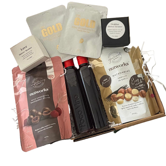 A curated gift box filled with gourmet snacks and packaged treats, photographed from above on a white surface. Visible items include Nutworks raspberry olive spread, Nutworks vanilla macadamia spread, two dark chocolate bars in clear bottles, two foil packets labeled GOLD, a small cream box, and a black round package.