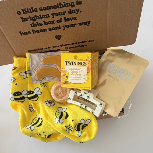 A curated gift box set containing a pair of bright yellow bee-printed socks, a box of Twinings Honey Honeybush tea, a gold honey pouch, and a clear packet of patchwork adhesive bandages, arranged inside an open brown cardboard box with printed text.