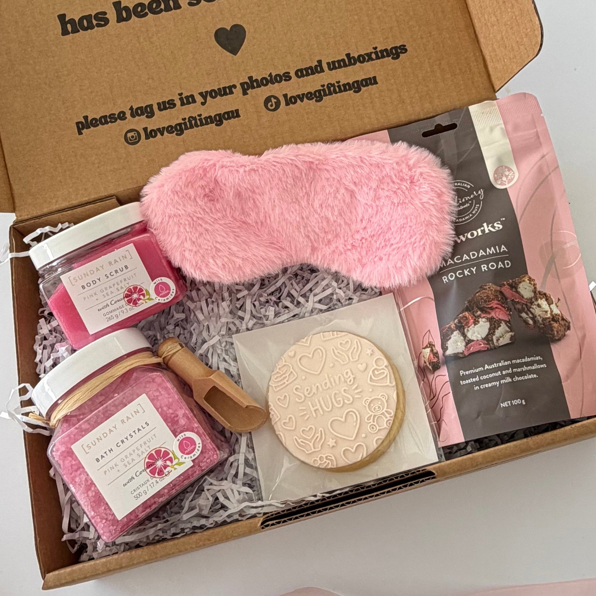 A curated gift box set containing a fluffy pink sleep mask, pink bath salts, a pink candle, a round embossed cookie, and a bag of chocolate from the brand 'love & works', arranged in shredded paper inside an open cardboard box.