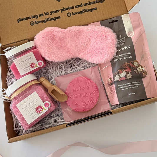 A curated gift box set on a white surface, featuring a fluffy pink sleep mask, pink bath salts, a pink soap bar, and a pink macadamia snack pouch inside an open brown cardboard box with shredded paper.