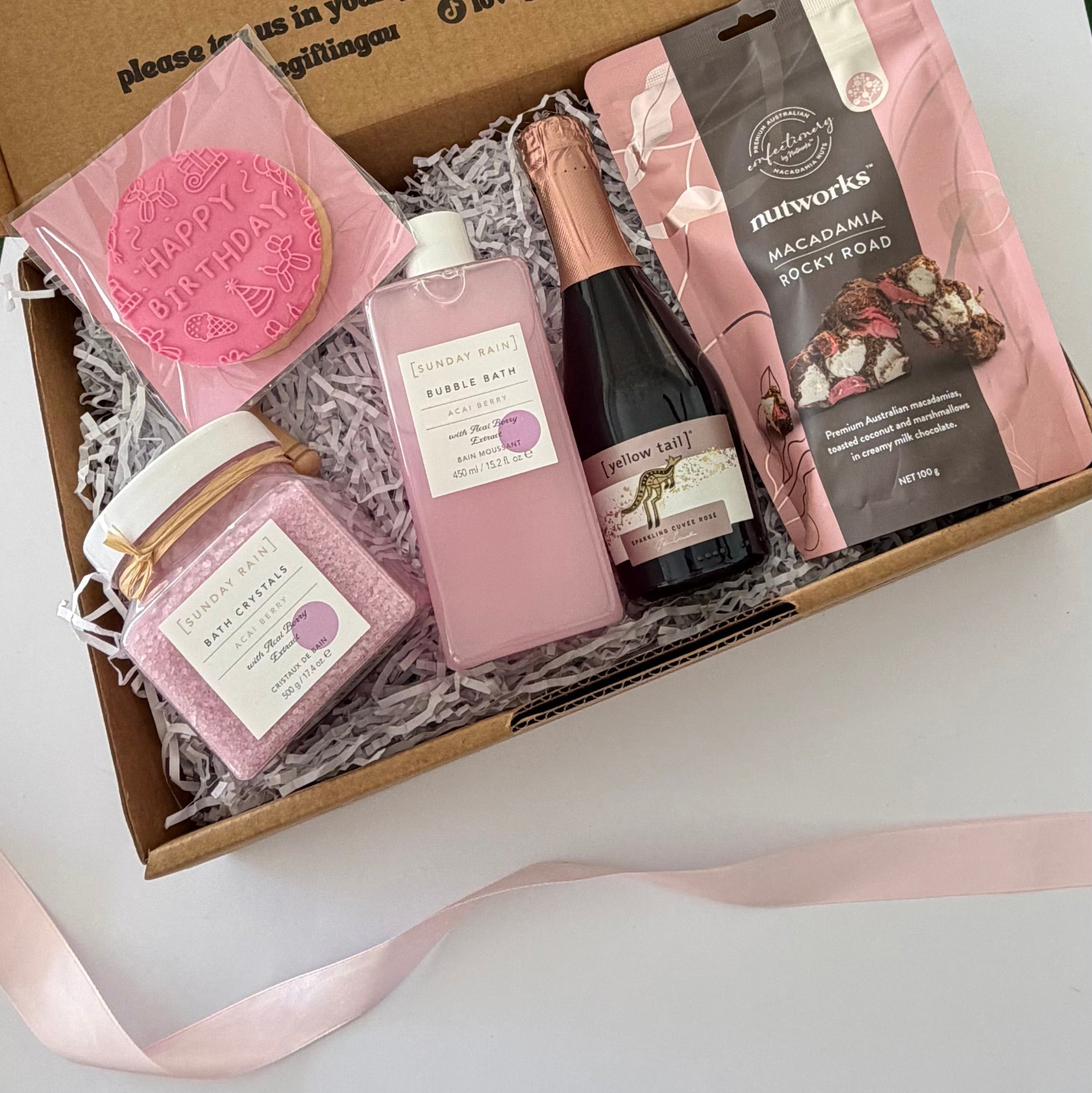 A curated gift box filled with pink-themed items, including a pink bath bomb, bubble bath, bath salts, a small bottle of sparkling wine, and a bag of Nutworks macadamia rocky road, arranged in white paper shred inside a cardboard box with a pink ribbon on a white surface.