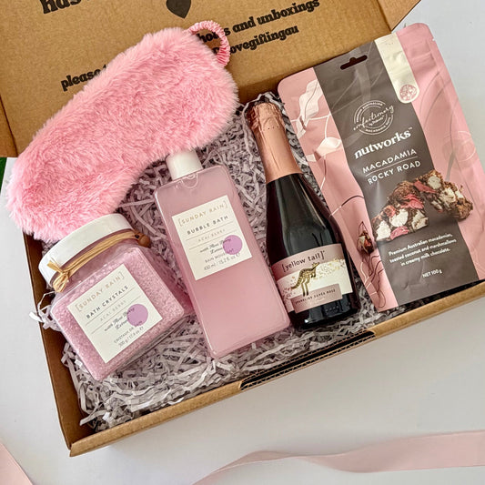 A curated gift box set containing a fluffy pink eye mask, a pink bubble bath bottle, a pink bath salt jar, a small bottle of prosecco, and a bag of Nutworks Macadamia Rocky Road, all arranged in shredded paper inside a brown cardboard box.