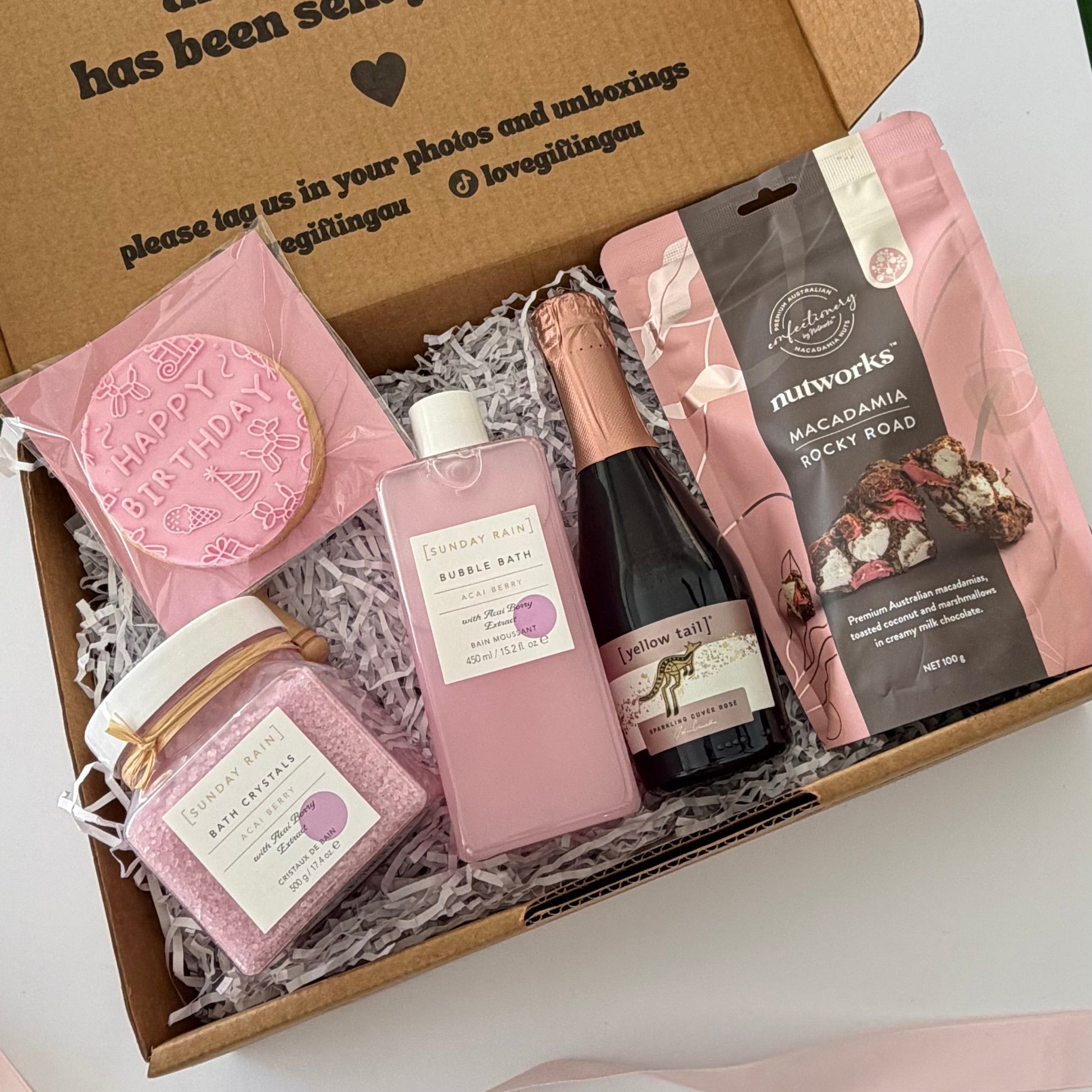 A curated gift box set containing a pink bath bomb, a jar of bath salts, a bottle of rosemary oil, a chocolate bar, and a pink soap bar, arranged in a brown cardboard box with shredded paper filler.