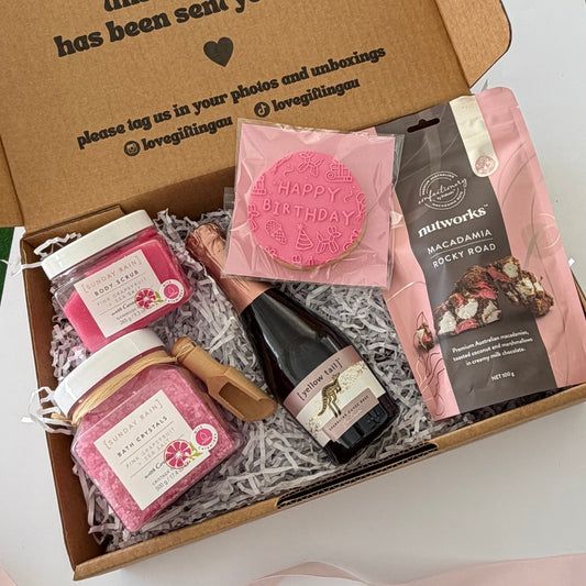 A curated gift box set displayed inside an open kraft cardboard box with printed text on the lid. The set includes pink bath salts in jars, a pink round candle, a small bottle of rosé wine, and a pink-and-black bag of macadamia powder, all arranged on white shredded paper with a pale pink ribbon around the box.
