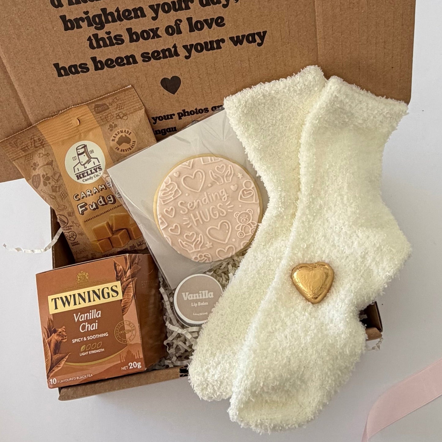 A curated gift box set containing a pair of cream fuzzy socks with a gold heart, a Twining's Vanilla Chai tea bag, a chocolate-covered cookie, and a small jar, arranged in a brown cardboard box with a message printed on the inside.