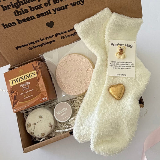 A curated gift box set featuring a cream-colored fluffy sock with a gold heart-shaped charm and a 'Pocket Hug' card, alongside a box of Twinings Vanilla Chai tea, a round heart-patterned soap, and a small round tin, all arranged inside an open brown cardboard box with printed text.