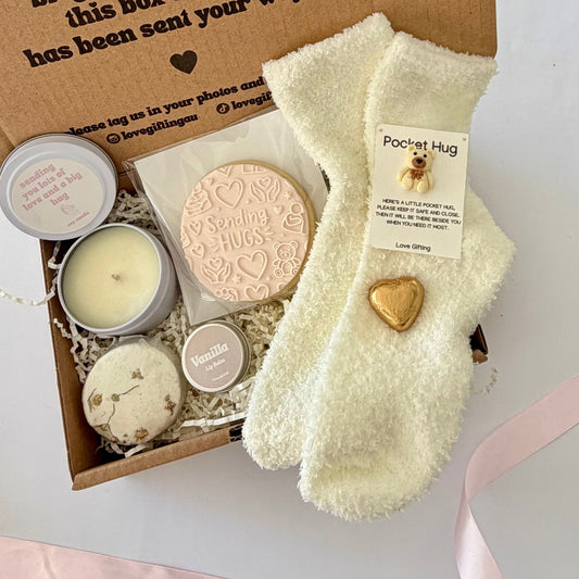 A cozy gift box filled with self-care and comfort items, including cream-colored fuzzy socks with a gold heart, a round bath bomb, a candle, and a candle tin, arranged in a flat-lay on a white background with a pink ribbon.