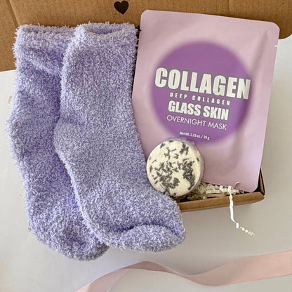A gift box set containing lavender fuzzy socks, a pink collagen glass skin overnight mask pouch, and a round herbal soap, arranged on a white surface with a pink ribbon.