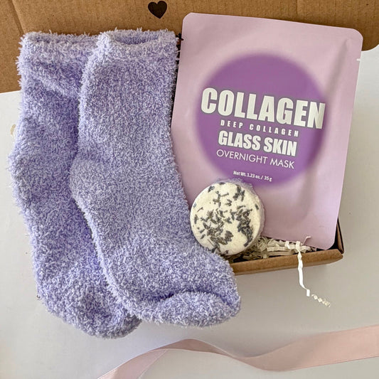 A gift box set containing lavender fuzzy socks, a pink collagen glass skin overnight mask pouch, and a round herbal soap, arranged on a white surface with a pink ribbon.