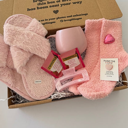 A cozy gift box filled with pink plush items, including fuzzy slippers, a pink mug, pink socks, two Twilly packs, a pink hair clip, and a small card reading 'Pocket rug LOVE'.
