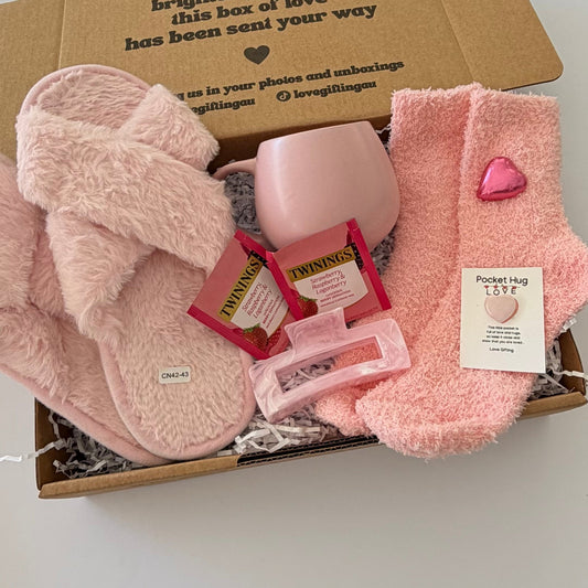A cozy gift box filled with pink plush items, including fuzzy slippers, a pink mug, pink socks, two Twilly packs, a pink hair clip, and a small card reading 'Pocket rug LOVE'.