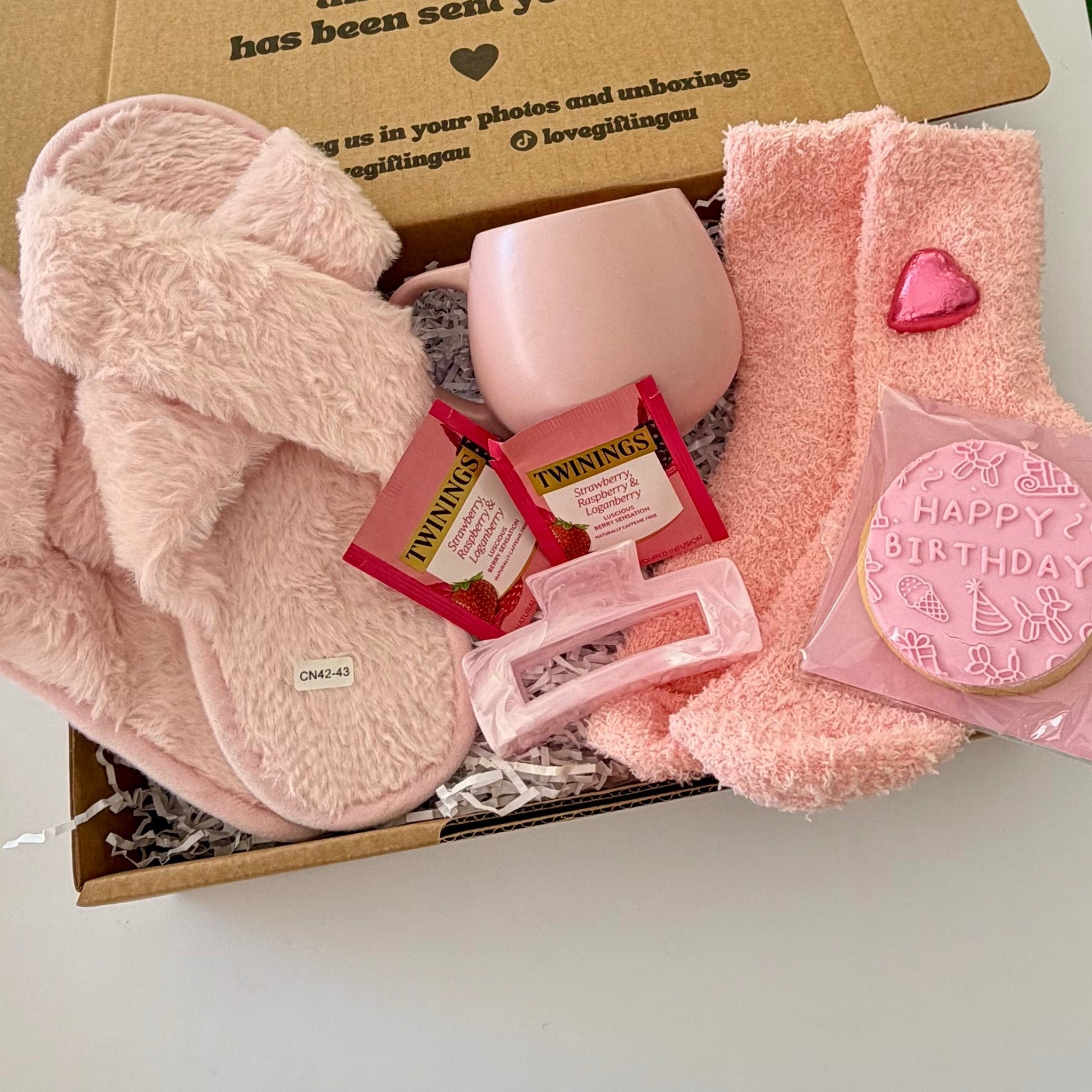 A pink-themed birthday gift box filled with plush pink slippers, a pink mug, a pink bath bomb, two packs of Twinings tea, a pink lip balm tube, and a pink 'Happy Birthday' cookie in a box printed with a warm message.
