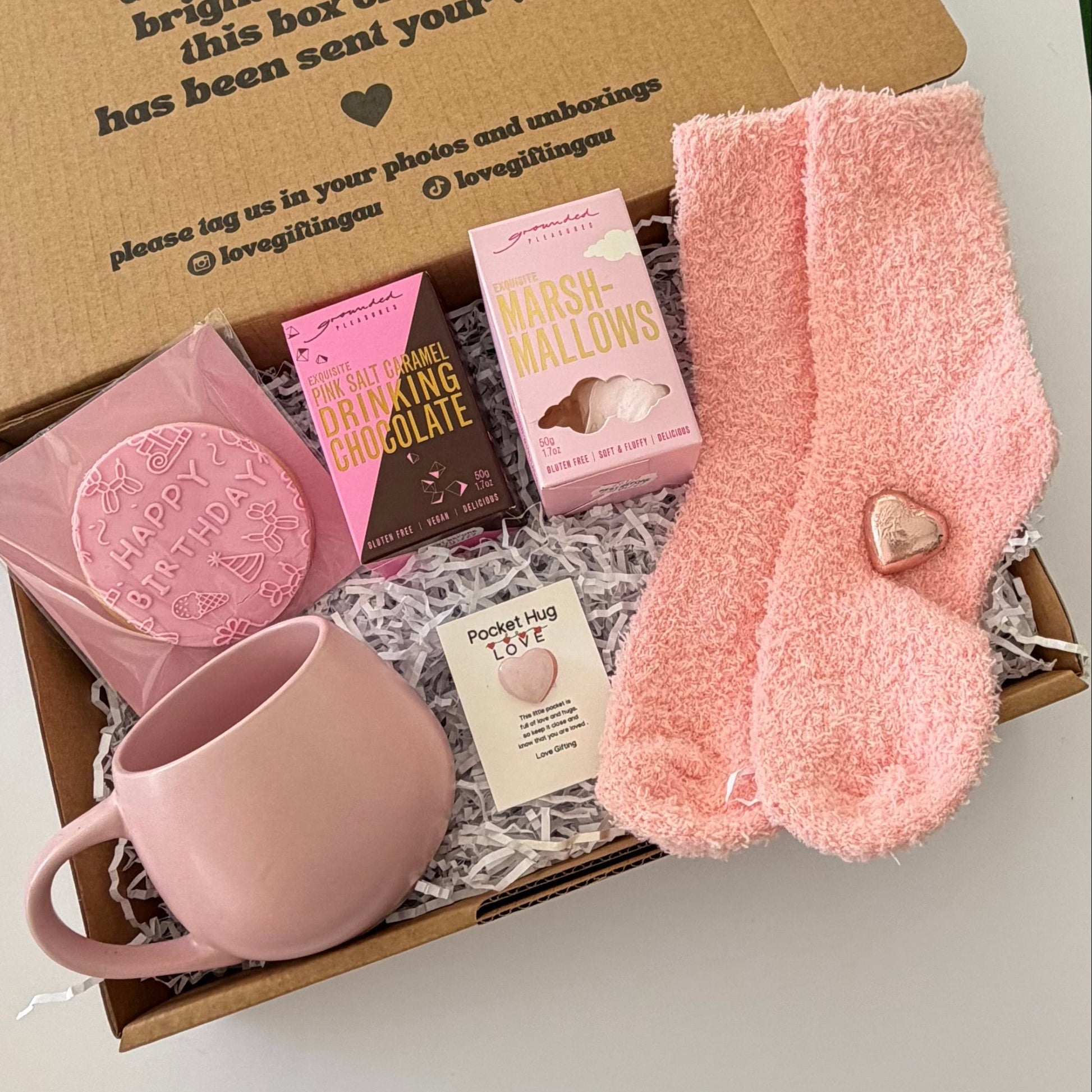 A cozy gift box filled with pink and brown treats, including a pink mug, fuzzy pink socks with a heart detail, a chocolate bar, marshmallows, and a bath bomb, arranged in shredded paper on a cardboard box.