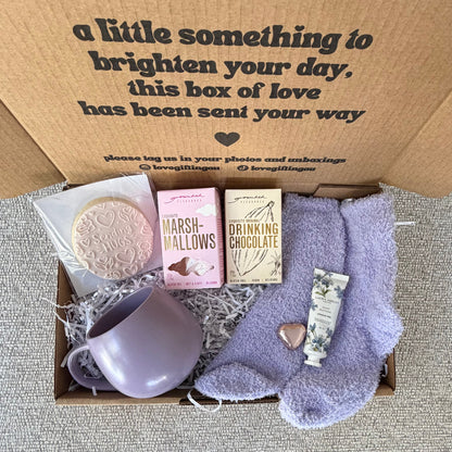 A curated gift box set featuring a lavender ceramic mug, lavender socks, a pink marshmallow cookie, a chocolate bar, and a small heart-shaped item, all arranged inside a brown cardboard box with printed text on the inside lid.
