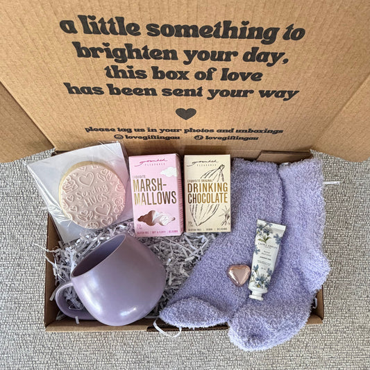 A curated gift box set featuring a lavender ceramic mug, lavender socks, a pink marshmallow cookie, a chocolate bar, and a small heart-shaped item, all arranged inside a brown cardboard box with printed text on the inside lid.